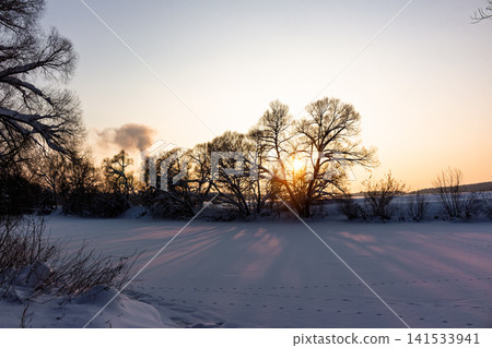 Golden hour paints a serene winter river landscape. Sun's vibrant light streams through bare trees, casting long shadows across the pristine snow. Pure natural beauty 141533941