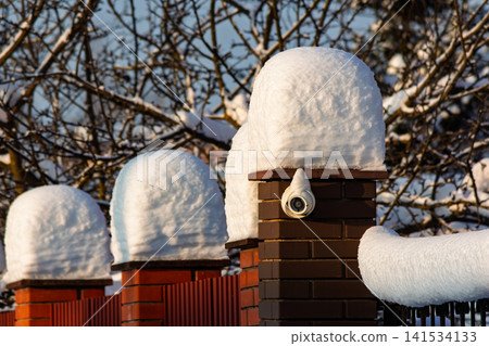 Winter security camera mounted on a brick pillar covered in snow, with snowy fence posts and bare trees in the background under sunlight Winter security camera mounted on a brick pillar covered in snow, with snowy fence posts and bare trees in the background under sunlight 141534133