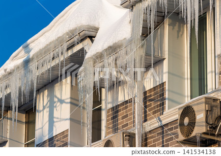 Brilliant, dagger-like icicles dangle from a snow-heavy roof under a vivid blue sky. Winter's icy peril and frosty charm illuminated by bright sun Brilliant, dagger-like icicles dangle from a snow-heavy roof under a vivid blue sky. Winter's icy peril and frosty charm illuminated by bright sun 141534138