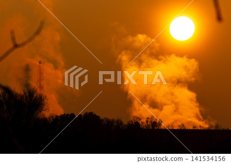 Blazing winter sun bathes thick industrial vapor in golden light. A tall smokestack casts a subtle shadow over the glowing steam, against a stunning orange sky Blazing winter sun bathes thick industrial vapor in golden light. A tall smokestack casts a subtle shadow over the glowing steam, against a stunning orange sky 141534156