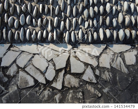 A gray stone wall with a rough, textured surface. Beneath it is a sloping slope, densely lined with rows of rounded, light-colored pebbles. The wall is combined with decorative paving. 141534479
