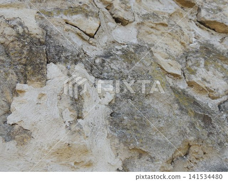 An old stone wall made of uneven beige-toned limestone. The rough, textured surface reveals cracks, dark stains, gray spots, traces of weathering, and mortar residue. 141534480