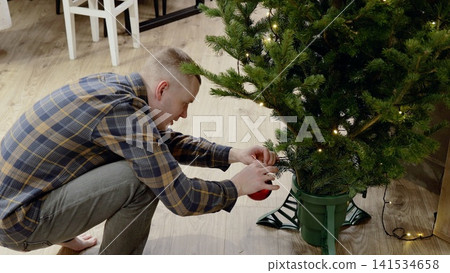 Man carefully hanging red christmas ball ornament on pre lit artificial tree while decorating home during festive season Man carefully hanging red christmas ball ornament on pre lit artificial tree while decorating home during festive season 141534658