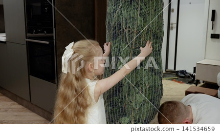 Young girl in white dress gently touching decorated christmas tree while father unwrapping holiday ornaments inside modern apartment Young girl in white dress gently touching decorated christmas tree while father unwrapping holiday ornaments inside modern apartment 141534659