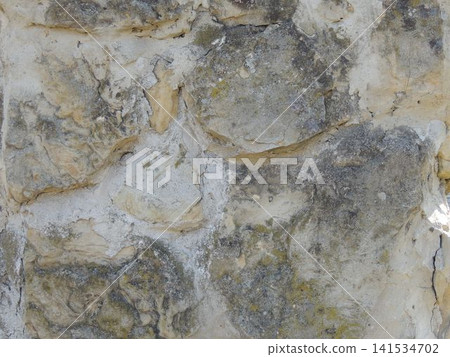 An old stone wall made of uneven beige and gray cobblestones. The rough, textured surface reveals traces of light-colored cement mortar, a dark layer of weathering, and patches of moss. An old stone wall made of uneven beige and gray cobblestones. The rough, textured surface reveals traces of light-colored cement mortar, a dark layer of weathering, and patches of moss. 141534702