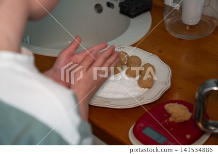 Child rolls cookie dough into small balls beside measured ingredients during homemade baking preparation in family kitchen. Childhood, home cooking, learning, parenting, family tradition, lifestyle 141534886