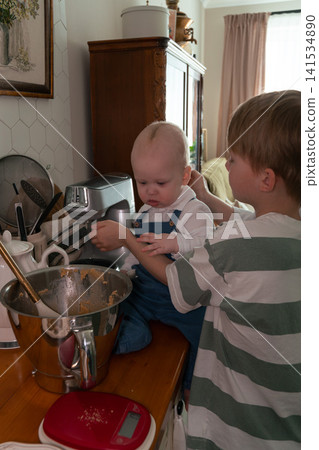 Older brother holds baby sibling beside bowl of homemade dough in family kitchen during baking preparation at home. Family, sibling bond, parenting, childhood, home cooking, lifestyle, togetherness Older brother holds baby sibling beside bowl of homemade dough in family kitchen during baking preparation at home. Family, sibling bond, parenting, childhood, home cooking, lifestyle, togetherness 141534890