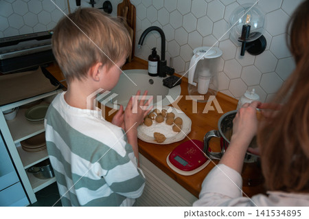 Child shapes cookie dough while adult prepares baking mixture in cozy home kitchen with homemade ingredients and family recipe. Childhood, home cooking, learning, parenting, lifestyle, family Child shapes cookie dough while adult prepares baking mixture in cozy home kitchen with homemade ingredients and family recipe. Childhood, home cooking, learning, parenting, lifestyle, family 141534895