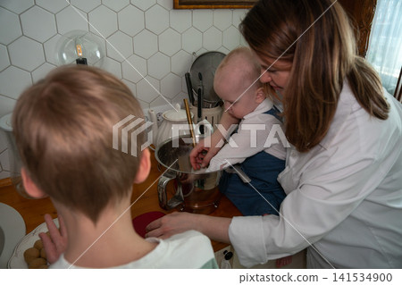 Mother helps baby stir homemade dough while older child joins family baking activity in cozy kitchen at home. Family, parenting, childhood, home cooking, bonding, education, lifestyle, togetherness Mother helps baby stir homemade dough while older child joins family baking activity in cozy kitchen at home. Family, parenting, childhood, home cooking, bonding, education, lifestyle, togetherness 141534900