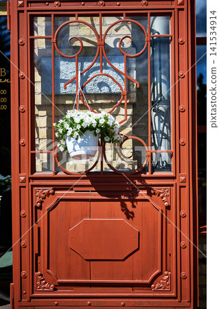 Hanging basket filled with white flowers sits on a decorative red metal door frame featuring classic iron patterns under bright sunlight during a warm summer afternoon Hanging basket filled with white flowers sits on a decorative red metal door frame featuring classic iron patterns under bright sunlight during a warm summer afternoon 141534914