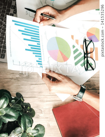 Businessperson reviews bar and pie charts at desk with plant Businessperson reviews bar and pie charts at desk with plant 141535296
