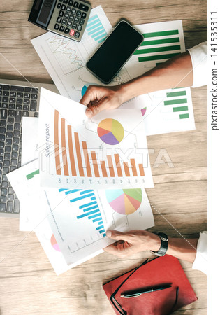 Top-down view of person reviewing business graphs and charts on wooden desk Top-down view of person reviewing business graphs and charts on wooden desk 141535311