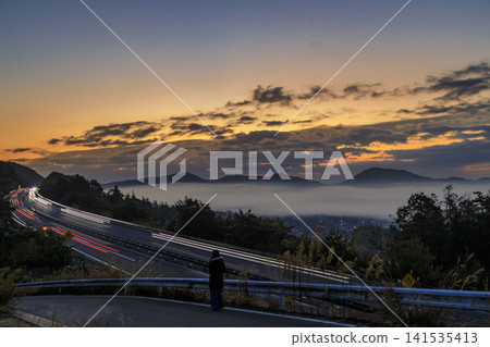 The dawn over a mountain village with a sea of clouds and the trails of cars on the highway. The dawn over a mountain village with a sea of clouds and the trails of cars on the highway. 141535413