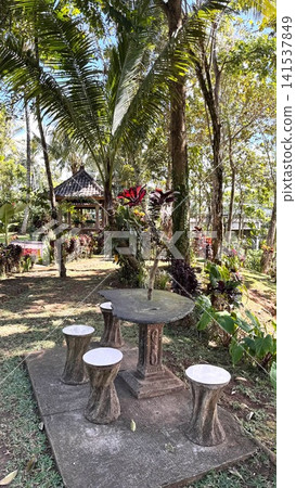 Relaxing outdoor seating area in a lush tropical garden with a stone table and stools. Surrounded by vibrant foliage and palm trees Relaxing outdoor seating area in a lush tropical garden with a stone table and stools. Surrounded by vibrant foliage and palm trees 141537849