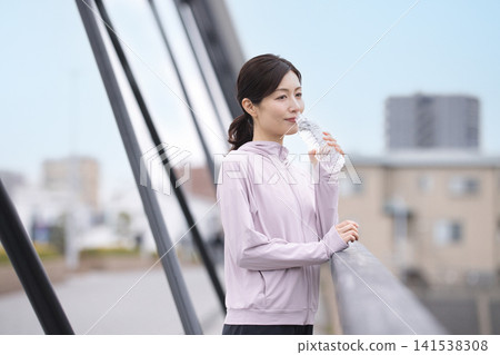 A middle-aged woman drinking bottled water while walking. A middle-aged woman drinking bottled water while walking. 141538308