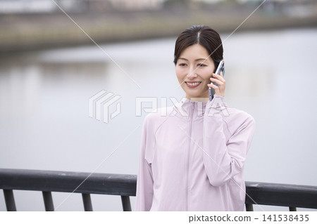 A middle-aged woman talking on her smartphone while walking in sportswear. A middle-aged woman talking on her smartphone while walking in sportswear. 141538435