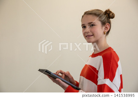 Girl in red striped sweater holding tablet, tapping touchscreen before beige wall, copy space Girl in red striped sweater holding tablet, tapping touchscreen before beige wall, copy space 141538995