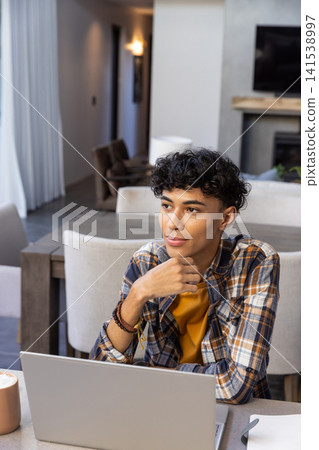 Asian man sitting at home dining area thinking beside silver laptop ceramic coffee mug notepad Asian man sitting at home dining area thinking beside silver laptop ceramic coffee mug notepad 141538997