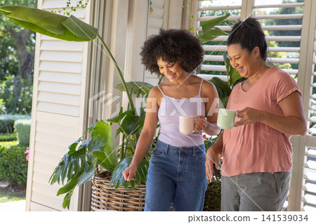 Diverse family holding pastel mugs, stepping on veranda with potted plant in basket and shutters 141539034