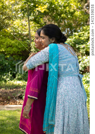 Indian mother and daughter embracing in sunlit garden, wearing fuchsia and turquoise outfits Indian mother and daughter embracing in sunlit garden, wearing fuchsia and turquoise outfits 141539038