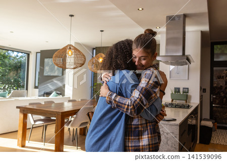 Diverse female friends hugging in modern kitchen with countertop island and rattan lights 141539096