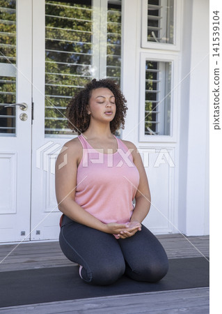 African American woman wearing pink top, leggings kneeling on yoga mat on wooden deck meditating 141539104