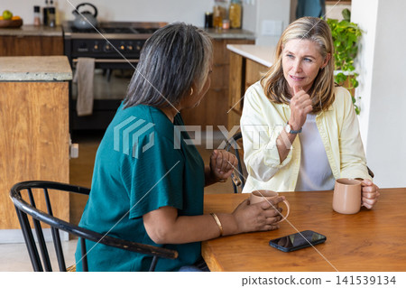 Senior female friends chatting while holding mugs at wooden kitchen table with smartphone Senior female friends chatting while holding mugs at wooden kitchen table with smartphone 141539134