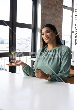 Mid adult woman wearing light green blouse, wristwatch gesturing at table in office, copy space Mid adult woman wearing light green blouse, wristwatch gesturing at table in office, copy space 141539152