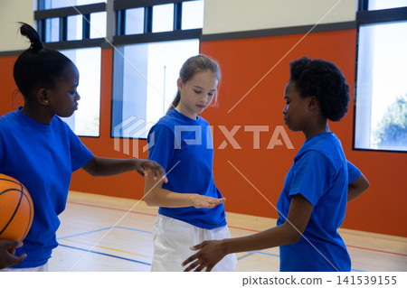 Diverse female children in blue shirts pointing at open palm while holding basketball on court Diverse female children in blue shirts pointing at open palm while holding basketball on court 141539155