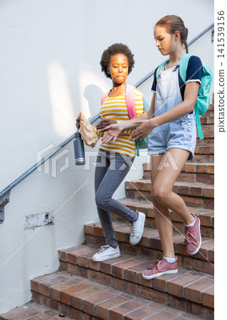 Diverse teenage girls with backpacks walking school steps holding bag, water bottle talking Diverse teenage girls with backpacks walking school steps holding bag, water bottle talking 141539156