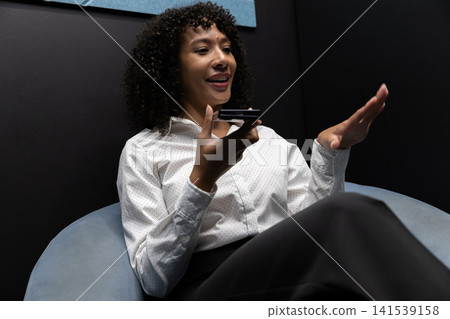 African american woman in business attire sitting in booth speaking into smartphone, gesturing African american woman in business attire sitting in booth speaking into smartphone, gesturing 141539158
