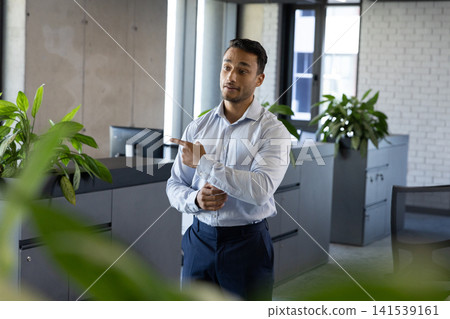 Asian man standing next to storage cabinets in modern office, gesturing with right hand 141539161