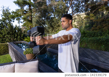 African american man lifting hex dumbbells on deck beside lap pool, sofa, plant, copy space African american man lifting hex dumbbells on deck beside lap pool, sofa, plant, copy space 141539179