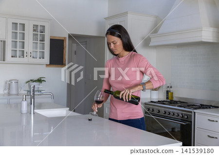 Woman pouring wine from green bottle into glass at kitchen island with sink, copy space Woman pouring wine from green bottle into glass at kitchen island with sink, copy space 141539180