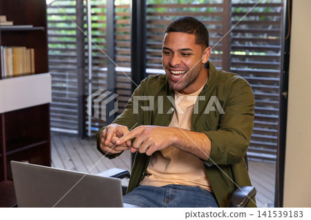 Man smiling and gesturing toward laptop screen in home office with wooden shelving, deck view 141539183