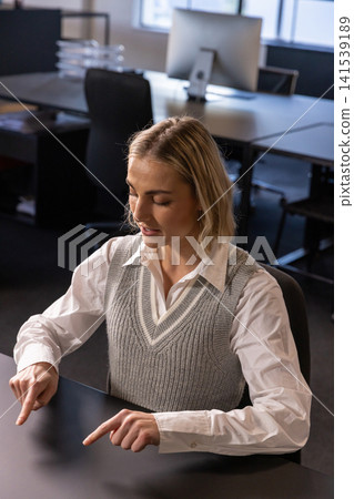 Woman pointing both index fingers downward at desk beside monitor in office workspace 141539189