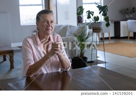 Woman sitting at wooden table in living room with neutral gray walls and potted plants Woman sitting at wooden table in living room with neutral gray walls and potted plants 141539201