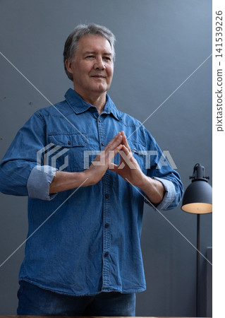 Senior man standing against grey wall in denim shirt making steeple gesture near black lamp Senior man standing against grey wall in denim shirt making steeple gesture near black lamp 141539226