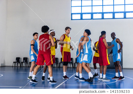 Diverse male basketball players in jerseys exchanging handshakes, hugs at gym court with chairs 141539233