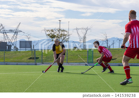 Teenage male athlete dribbling white field hockey ball on turf pitch holding red-black stick Teenage male athlete dribbling white field hockey ball on turf pitch holding red-black stick 141539238