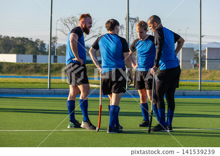 Diverse male teammates huddling on turf near goal discussing strategy with hockey sticks 141539239