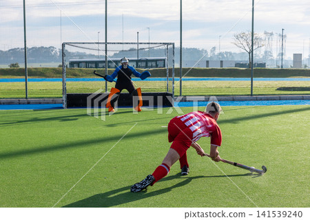 Teenage attacker lunging with stick on synthetic turf, goalkeeper diving with helmet Teenage attacker lunging with stick on synthetic turf, goalkeeper diving with helmet 141539240