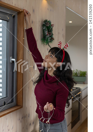 Woman wearing antler headband stretching upward at kitchen window hanging string lights Woman wearing antler headband stretching upward at kitchen window hanging string lights 141539260