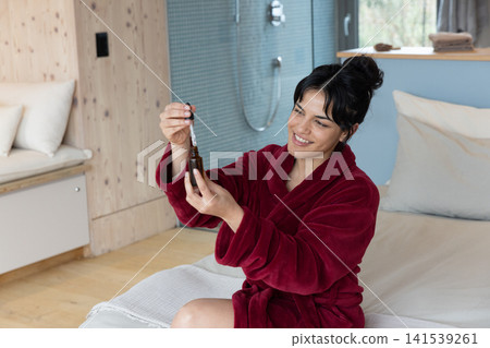 Asian woman sitting in bathrobe examining brown dropper bottle on bed in bedroom, copy space Asian woman sitting in bathrobe examining brown dropper bottle on bed in bedroom, copy space 141539261
