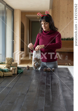 Hispanic adult woman wearing headband placing pinecones into vase with gifts on table Hispanic adult woman wearing headband placing pinecones into vase with gifts on table 141539262
