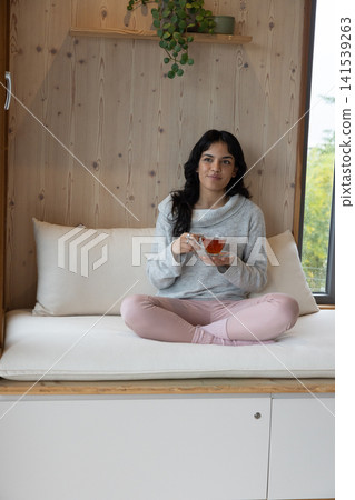Asian female sitting on bench in wood-paneled home nook sipping tea by plant shelf Asian female sitting on bench in wood-paneled home nook sipping tea by plant shelf 141539263
