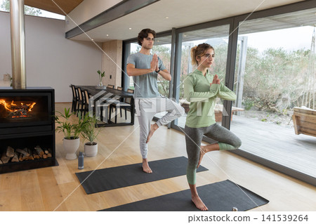 Couple practicing yoga pose in athletic wear on black mats in living area with wood stove 141539264