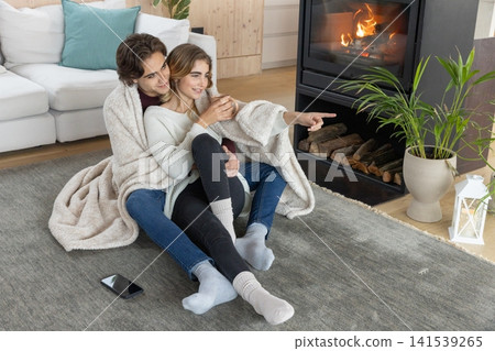 Couple snuggling together in cream blanket on gray rug in front of fireplace holding white mug Couple snuggling together in cream blanket on gray rug in front of fireplace holding white mug 141539265
