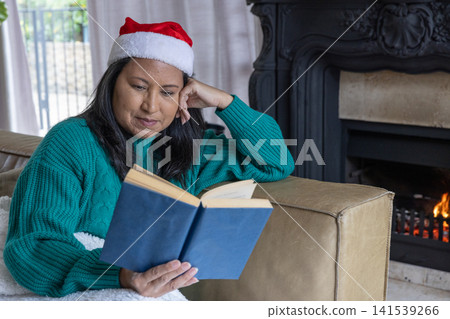 Asian woman sitting in living room on couch in santa hat reading blue book with fireplace Asian woman sitting in living room on couch in santa hat reading blue book with fireplace 141539266