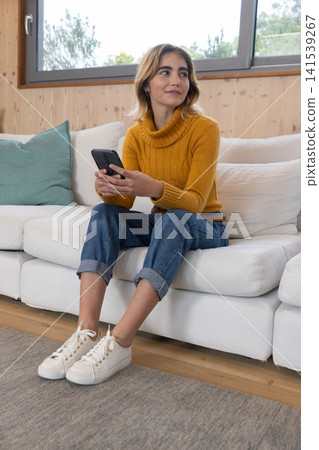 Woman wearing mustard sweater holding smartphone while sitting on white couch in home living room Woman wearing mustard sweater holding smartphone while sitting on white couch in home living room 141539267